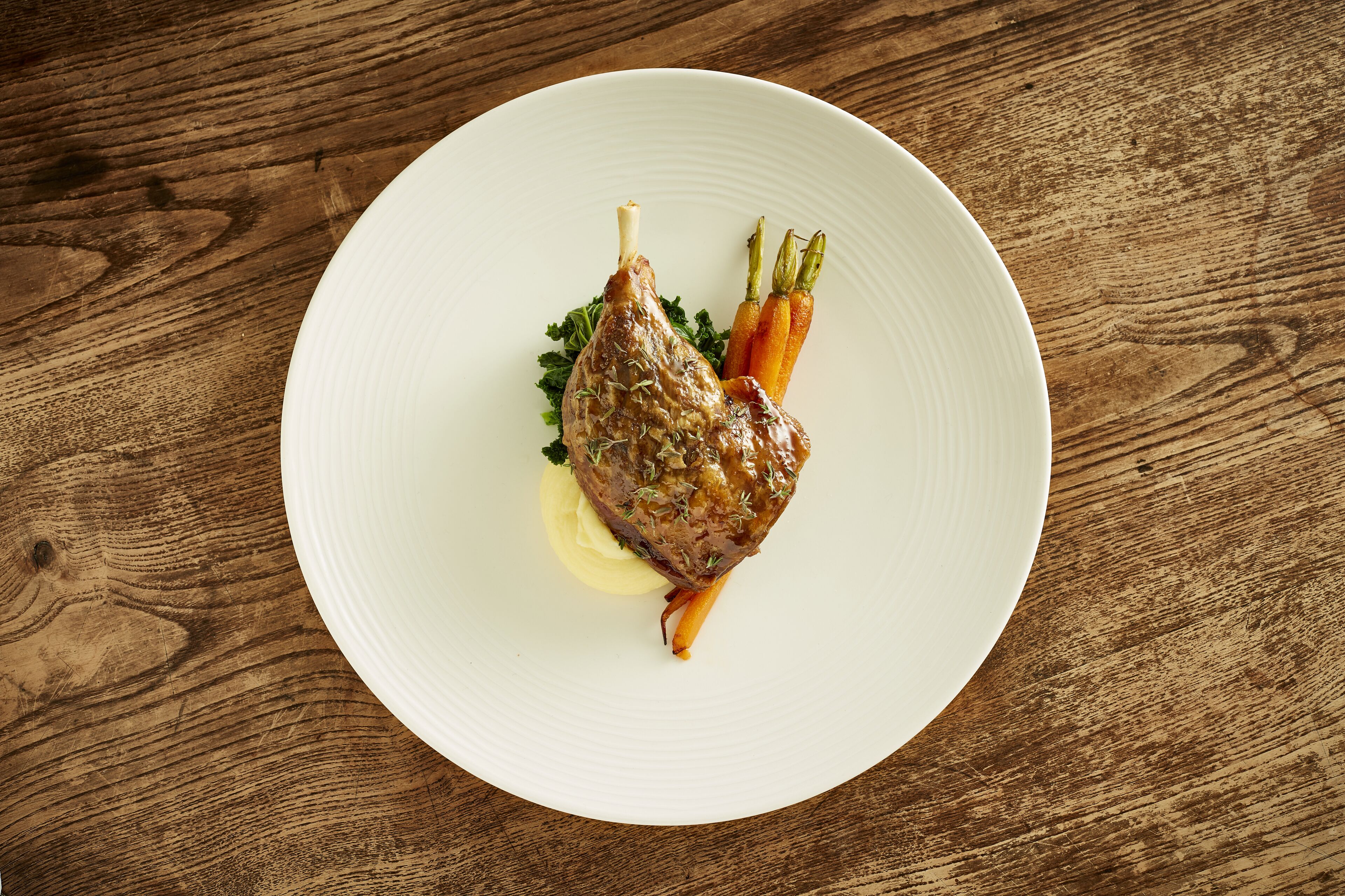 A white plate with a roasted lamb leg, carrots, and a garnish on a wooden table.