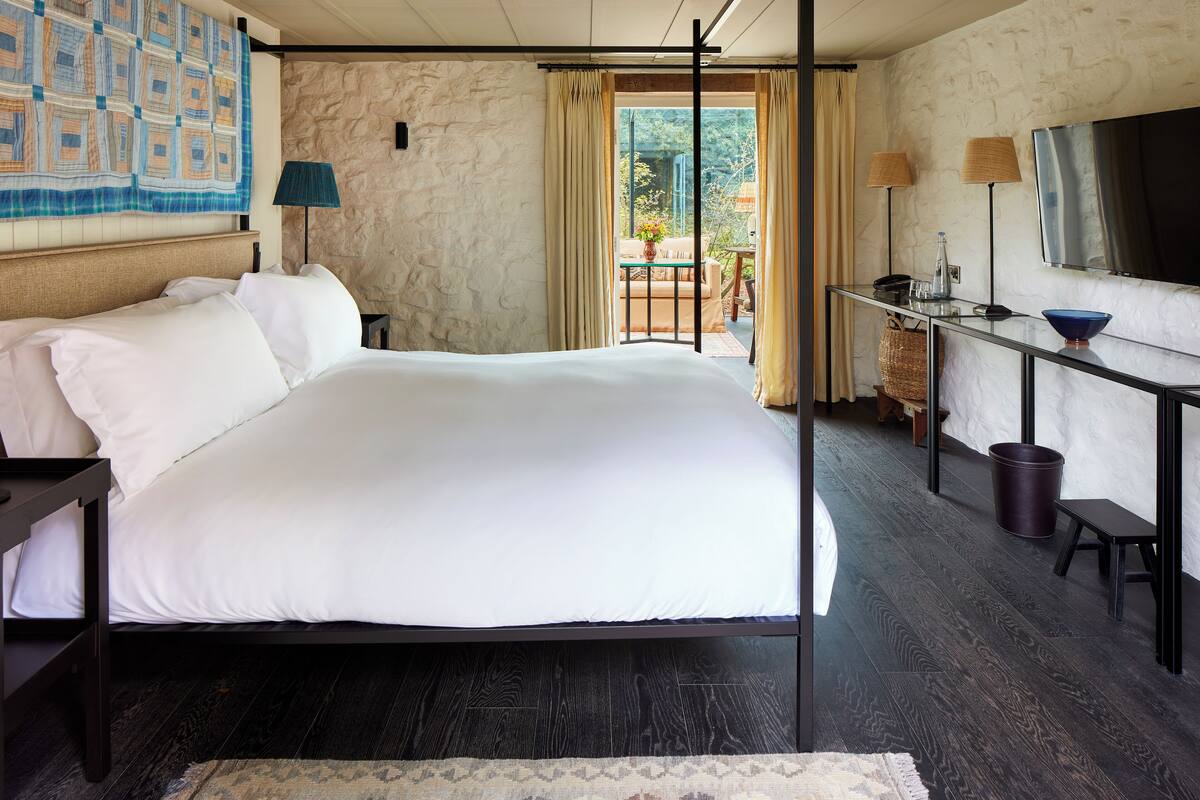 A modern hotel room with a large bed, a desk with a TV, and a balcony with a view of greenery.