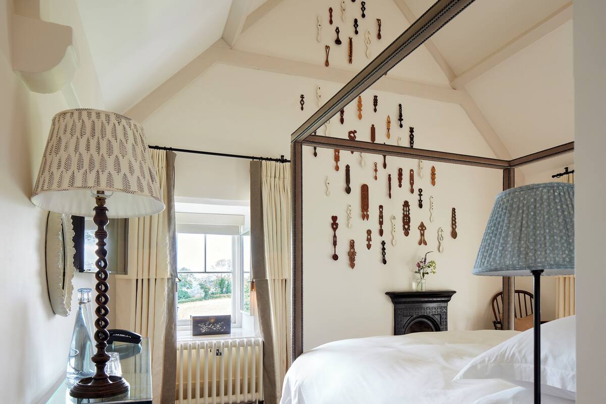 A bedroom with a four-poster bed, a bedside table with a lamp, a nightstand with books, and a window with curtains.