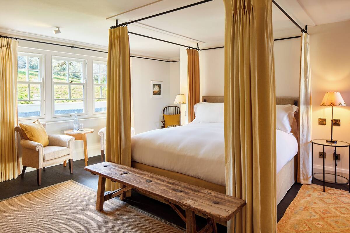 A four-poster bed with a wooden bench in front, a small round table, and a chair with a yellow cushion.