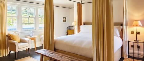 Una cama con dosel sobre una base de madera, con un banco de madera en frente, una mesita redonda pequeña y una silla con cojín amarillo.