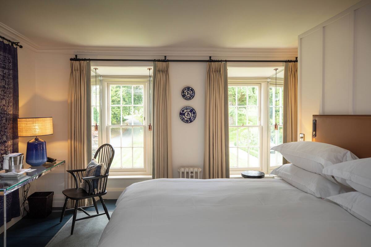 A hotel room with a large bed, a desk with a lamp, a chair, and two windows with curtains.