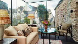Un espacio exterior acogedor con mesa de vidrio, sofá y sillón, con una pared de piedra y vistas a casas al fondo.