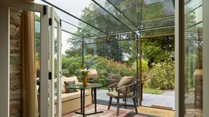 Un patio con paredes de vidrio, que incluye un sofá, una silla y una mesa, con vistas a un jardín y árboles.