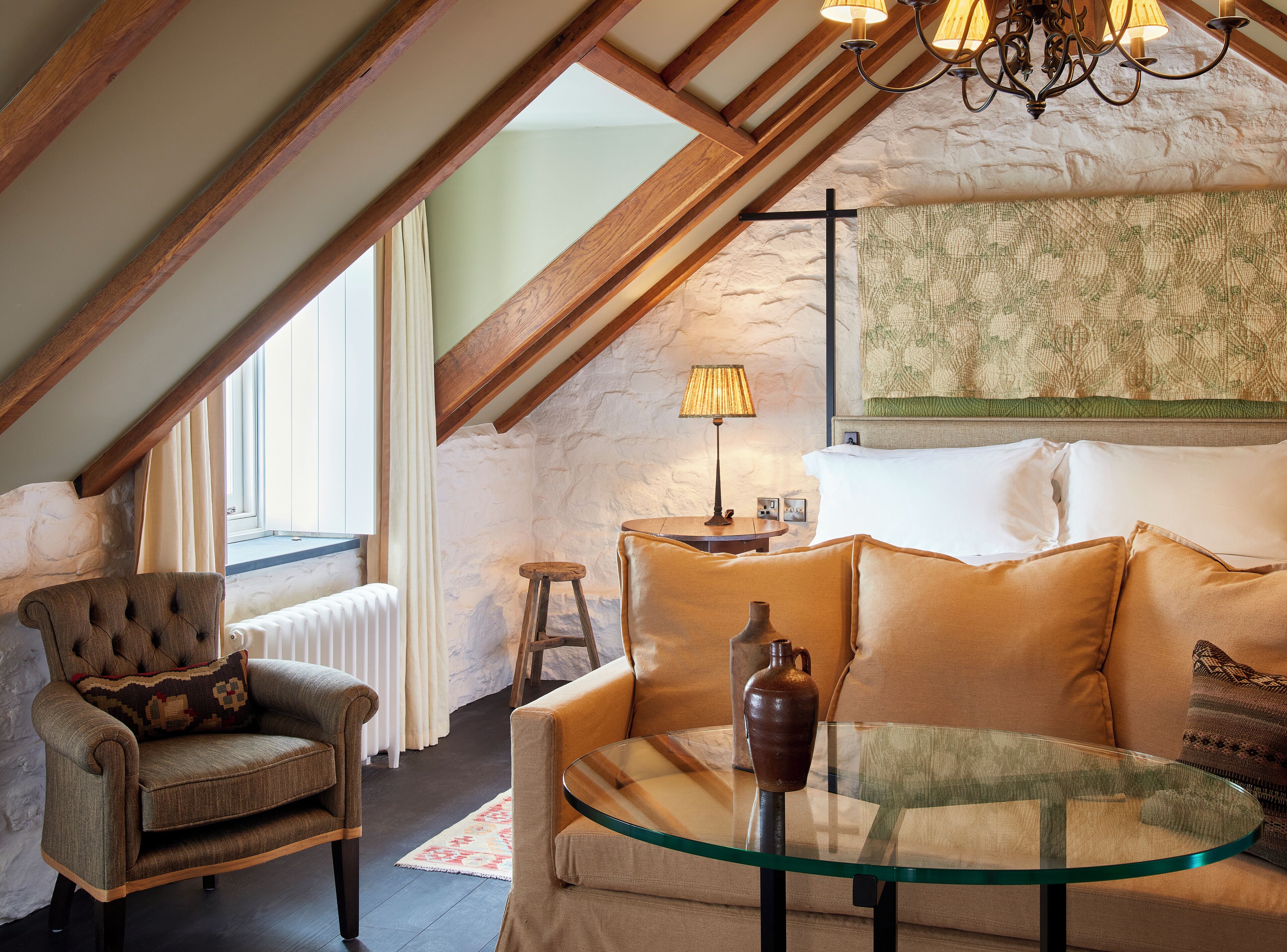 A cozy loft bedroom with a sloped ceiling, a wooden beam structure, a glass table, a sofa, and a chair.