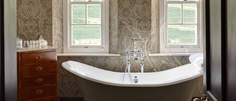 Ein Badezimmer mit freistehender Badewanne, zwei Fenstern mit Vorhängen, einem Waschtisch mit Schubladen und einer Tür.