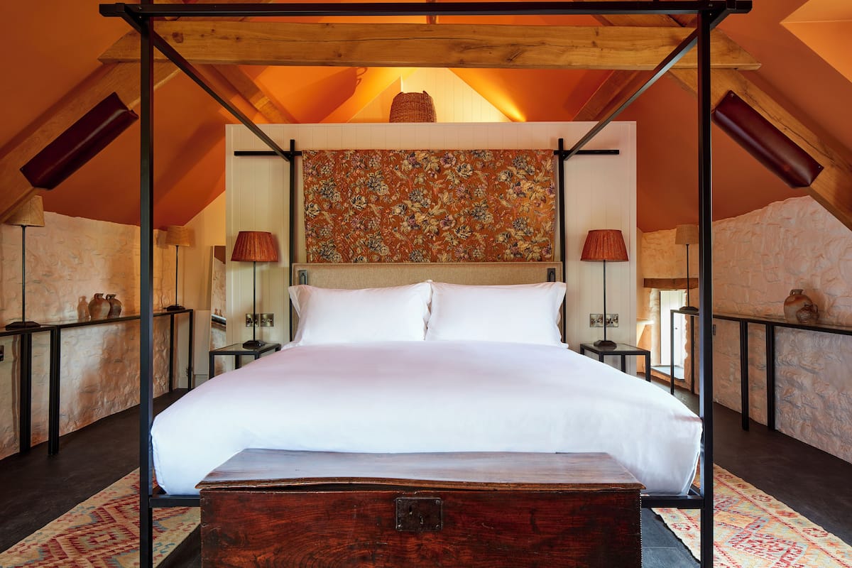 A four-poster bed with a wooden headboard, a large floral tapestry, and a stone wall backdrop.