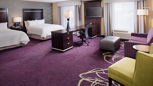 A hotel room with two beds, a desk, a chair, an ottoman, a flat-screen TV, and a large window with curtains.