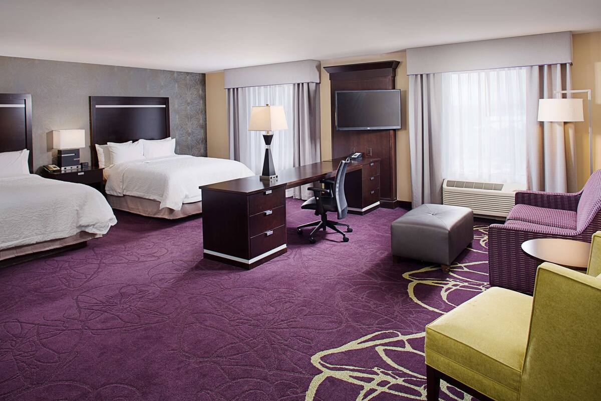 Une chambre d’hôtel avec deux lits, un bureau, une chaise, un pouf, un téléviseur à écran plat et une grande fenêtre avec des rideaux.