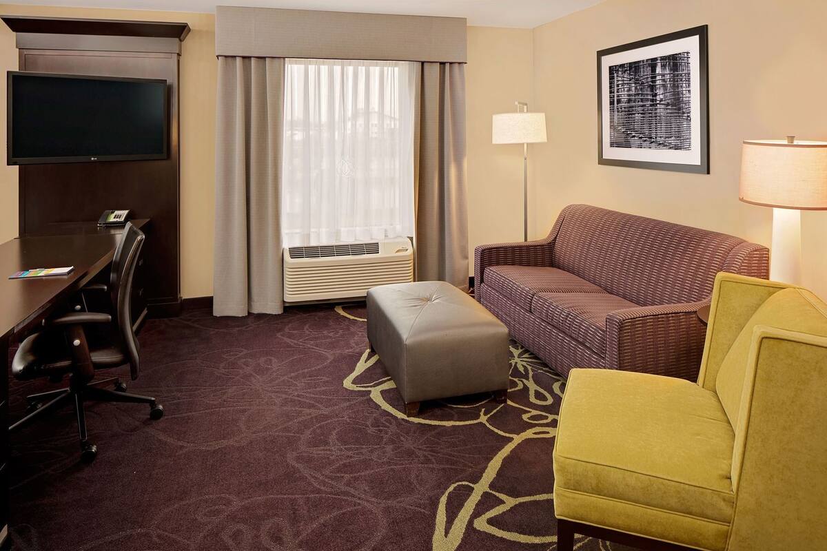 Une chambre d’hôtel équipée d’un téléviseur à écran plat, d’un bureau, d’un canapé, d’un pouf, d’une lampe et d’un tableau encadré au mur.