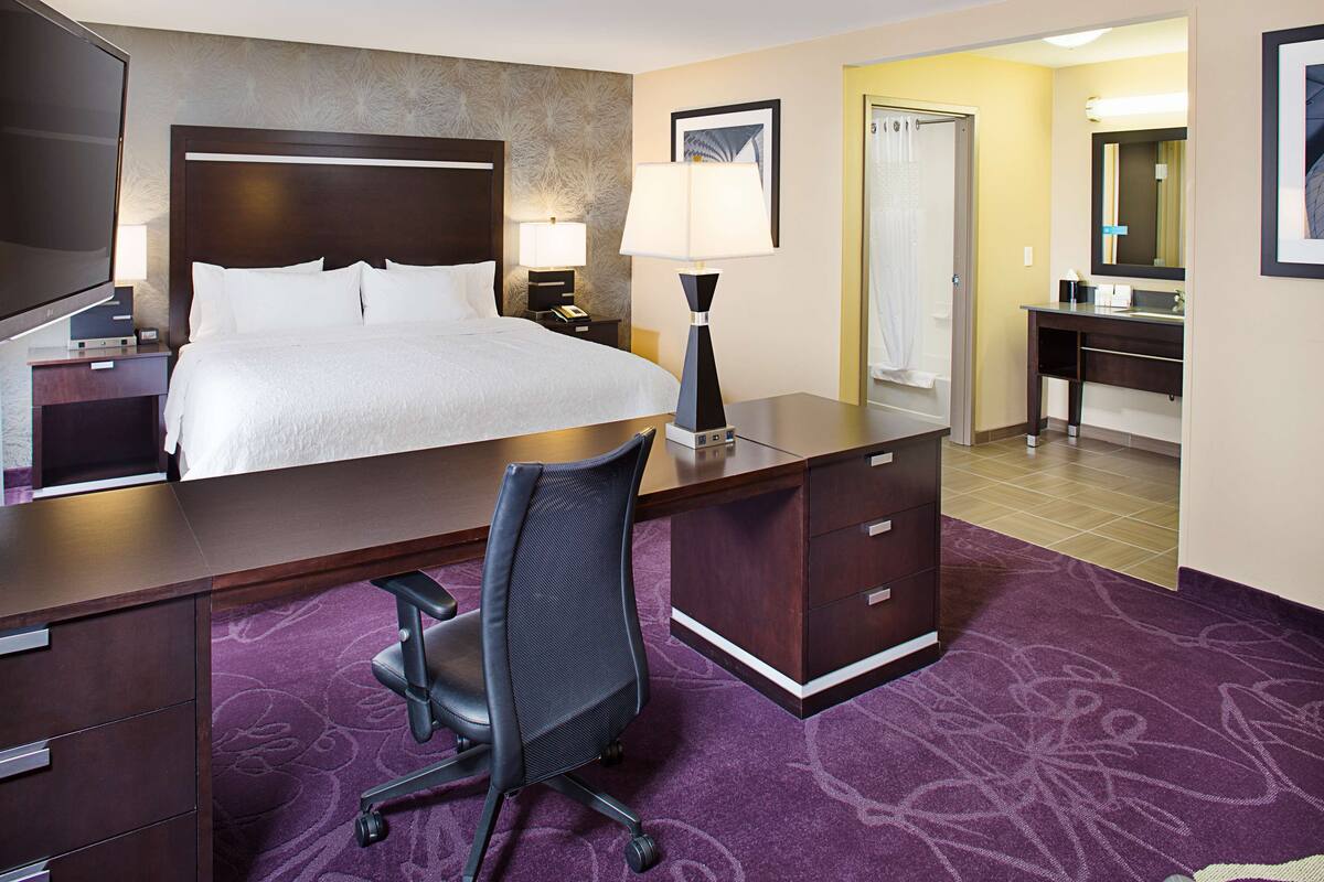 Une chambre d’hôtel avec un grand lit, un bureau avec une chaise, une lampe de bureau, une télévision et une salle de bain avec une douche et un miroir.