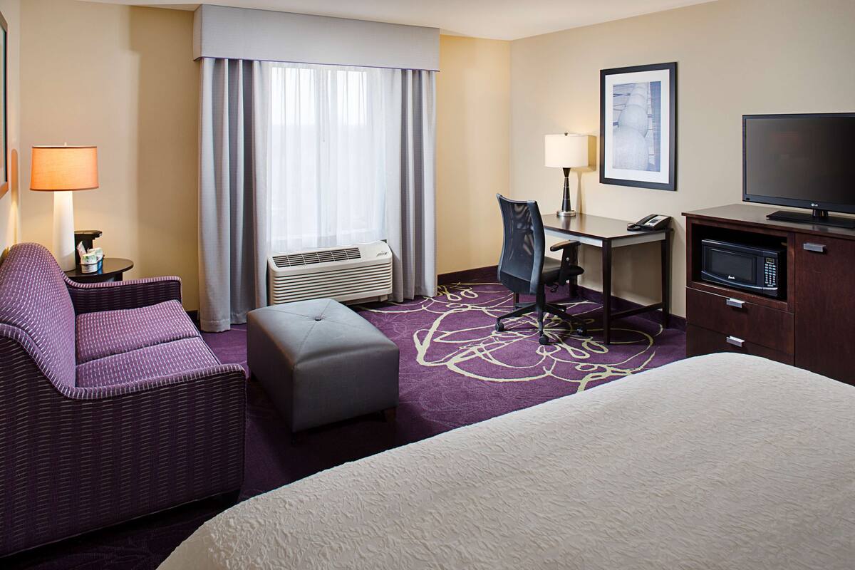 Une chambre d’hôtel équipée d’un lit, d’un bureau, d’une chaise, d’une télévision, d’un four à micro-ondes et d’une fenêtre avec des rideaux.