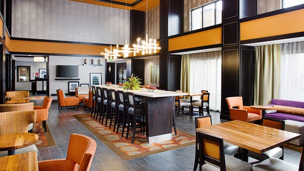 A modern hotel lobby with a central bar area, multiple seating options including sofas and chairs, and a large television screen.