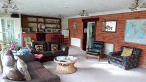A spacious living room with a leather sofa, a patterned armchair, and a coffee table. A brick wall with a world map and a bookshelf filled with books and decorative items are visible. - Bartles Lodge (Dereham)
