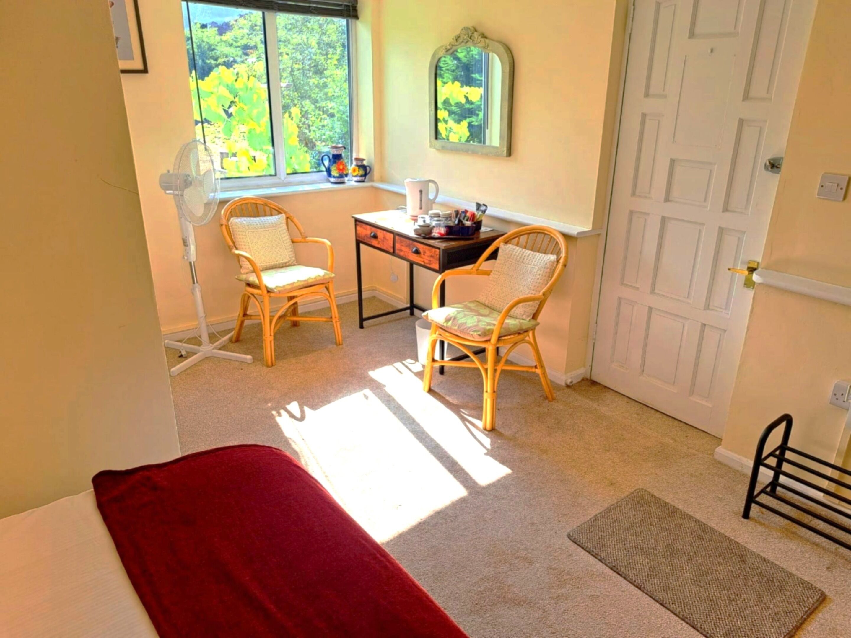 A room with a bed, two chairs, a desk, a window with a view of trees, and a door.