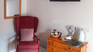 A room with a red chair, a wooden dresser with a basket and a kettle, a mirror, and a flat-screen TV on the wall. - Bartles Lodge (Dereham)