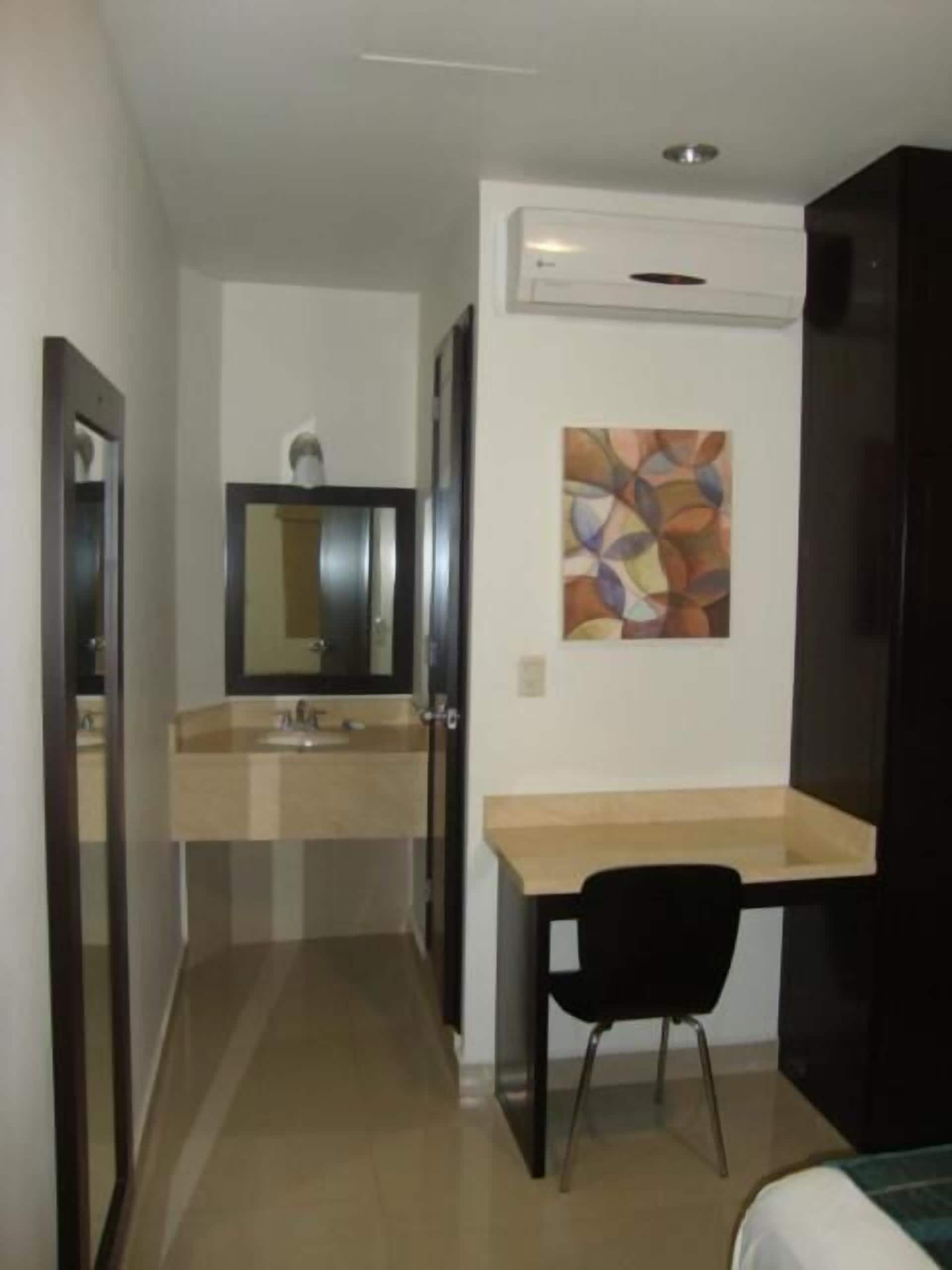 Une petite chambre moderne, équipée d’un lit, d’un bureau avec une chaise, d’un miroir et d’un climatiseur fixé au mur.