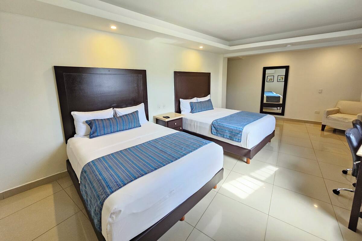 Habitación de hotel con dos camas, cada una con cabecera oscura, ropa de cama blanca y colchas a rayas azules. Hay una mesita de noche con lámpara, una silla y un espejo grande en la pared.