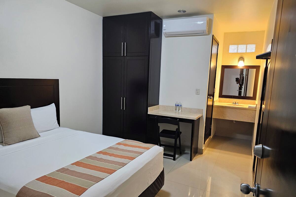 Habitación de hotel con cama, escritorio, armario, aire acondicionado, baño con espejo y puerta.