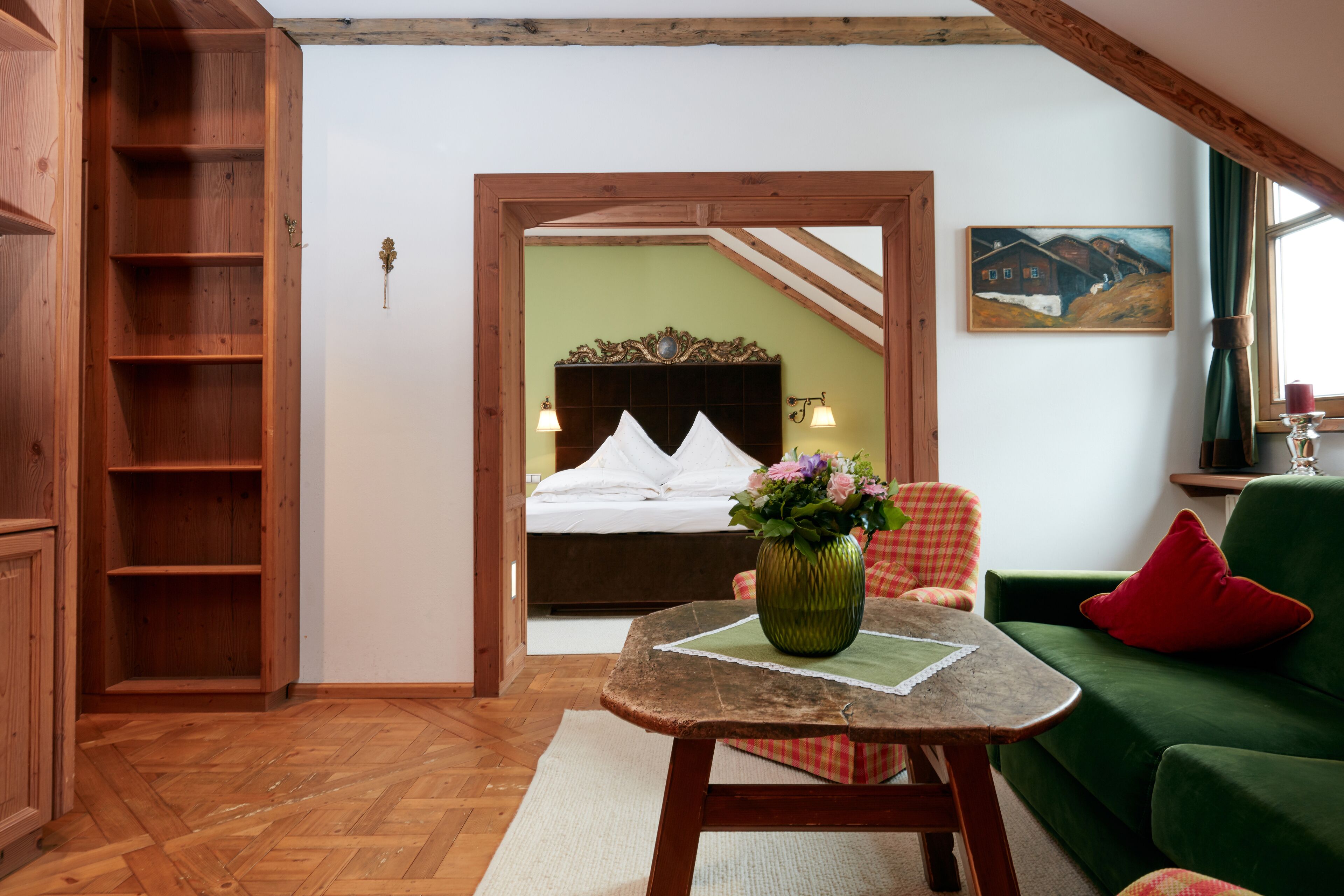 Ein Zimmer mit Holzboden, einem grünen Sofa, einem Holztisch mit einer Vase, einem Bett mit Kopfteil und einem Bild an der Wand.