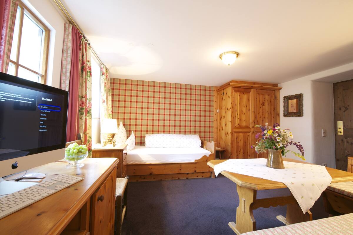 Habitación de hotel con cama de madera, un escritorio con computadora, una mesita con un florero y una pared a cuadros.