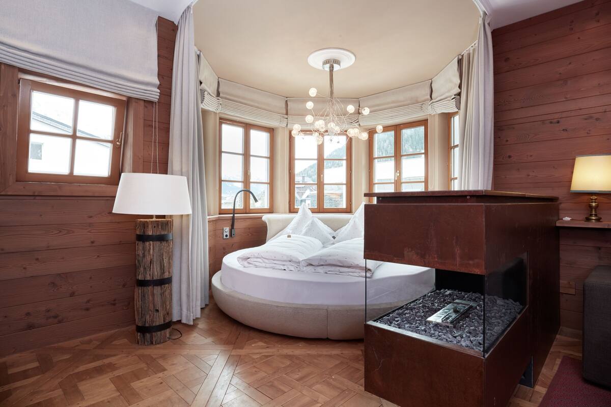 Un dormitorio acogedor con una cama redonda, una chimenea y grandes ventanales que ofrecen vistas al exterior.