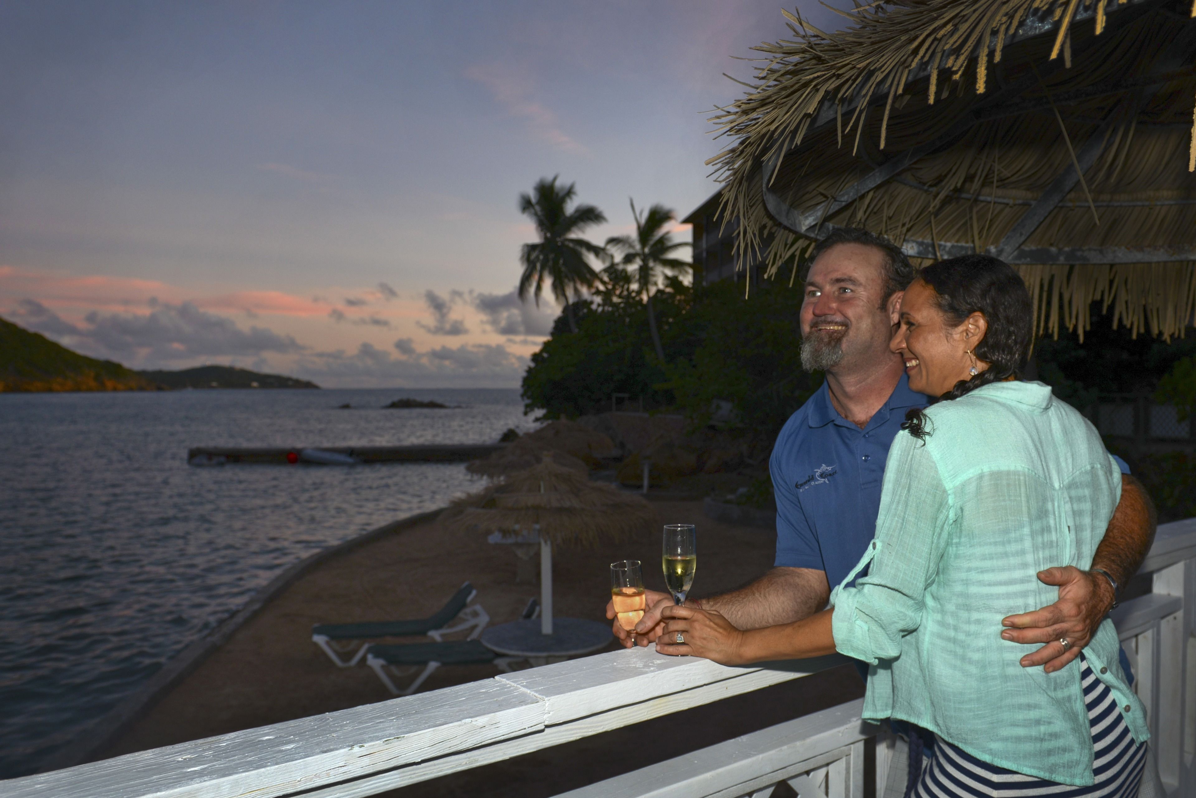 A couple on a balcony overlooking a beach with palm trees and a thatched-roof structure.
