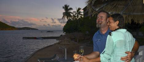 A couple on a balcony overlooking a beach with palm trees and a thatched-roof structure.