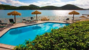 A swimming pool with lounge chairs and umbrellas, surrounded by lush greenery and a view of a lake with mountains in the background. - Lindbergh Bay Hotel and Villas (St. Thomas)