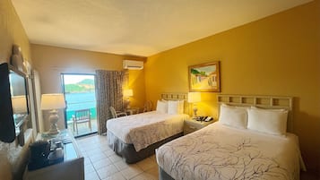 Une chambre d’hôtel avec deux lits, un bureau avec une lampe, une chaise, une petite table et une vue sur la mer.