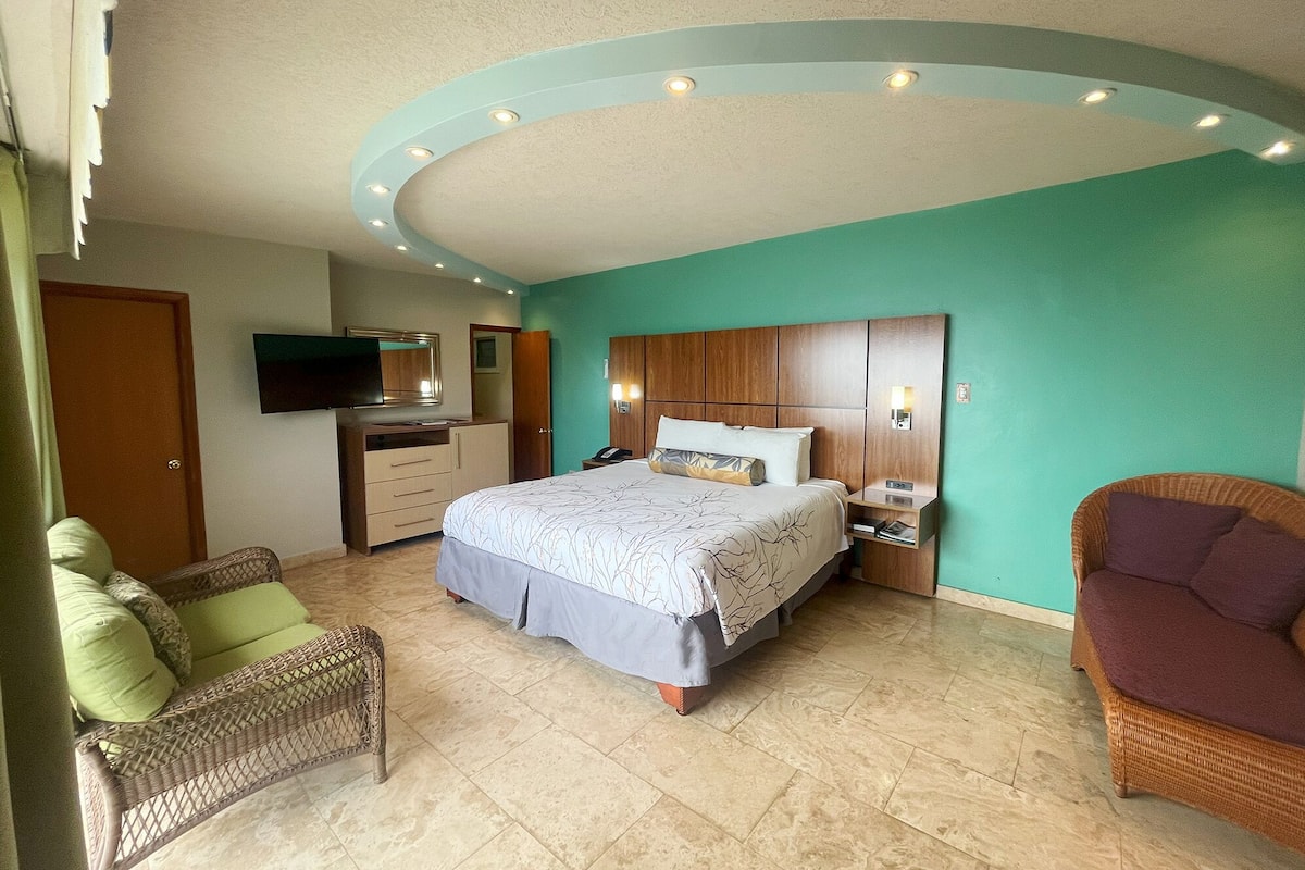 Ein Schlafzimmer mit einem Bett, einem Fernseher, einem Kleiderschrank, einem Nachttisch und zwei Korbstühlen.