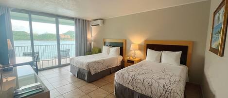 A hotel room with two beds, a desk, and a balcony with a view of the sea.