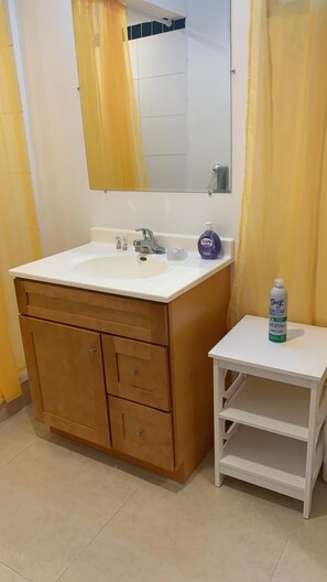 A bathroom with a white sink, wooden vanity, a mirror, and a small white shelf with a bottle of disinfectant. - The Horny Toad Guesthouse (Simpson Bay)