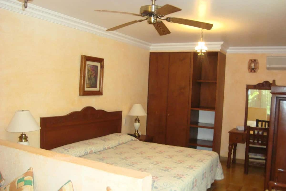 Un dormitorio con una cama, dos lámparas de noche, un ventilador de techo, un armario de madera, una mesita pequeña y una silla.