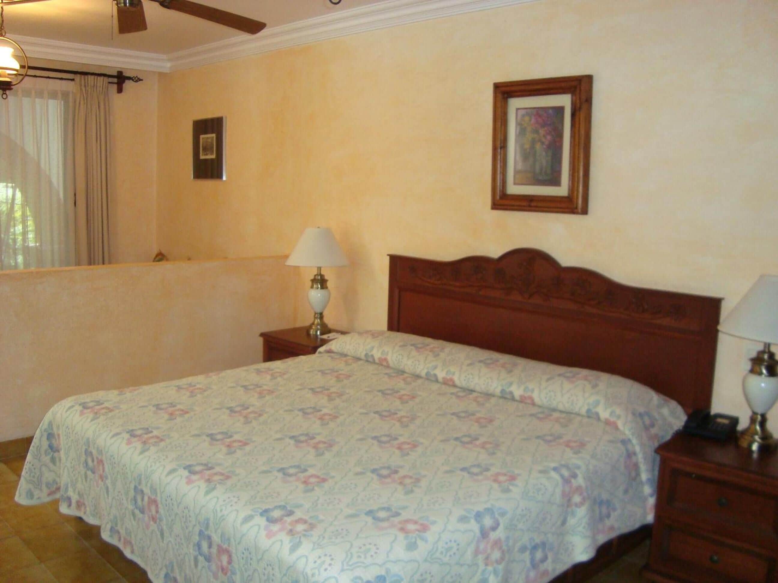 A bedroom with a wooden bed, floral bedspread, two bedside lamps, a framed picture on the wall, and a window with curtains.