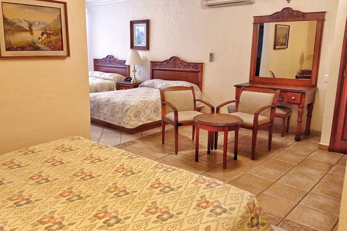 Una habitación de hotel con una cama, dos sillas, una mesita, una lámpara y un espejo.