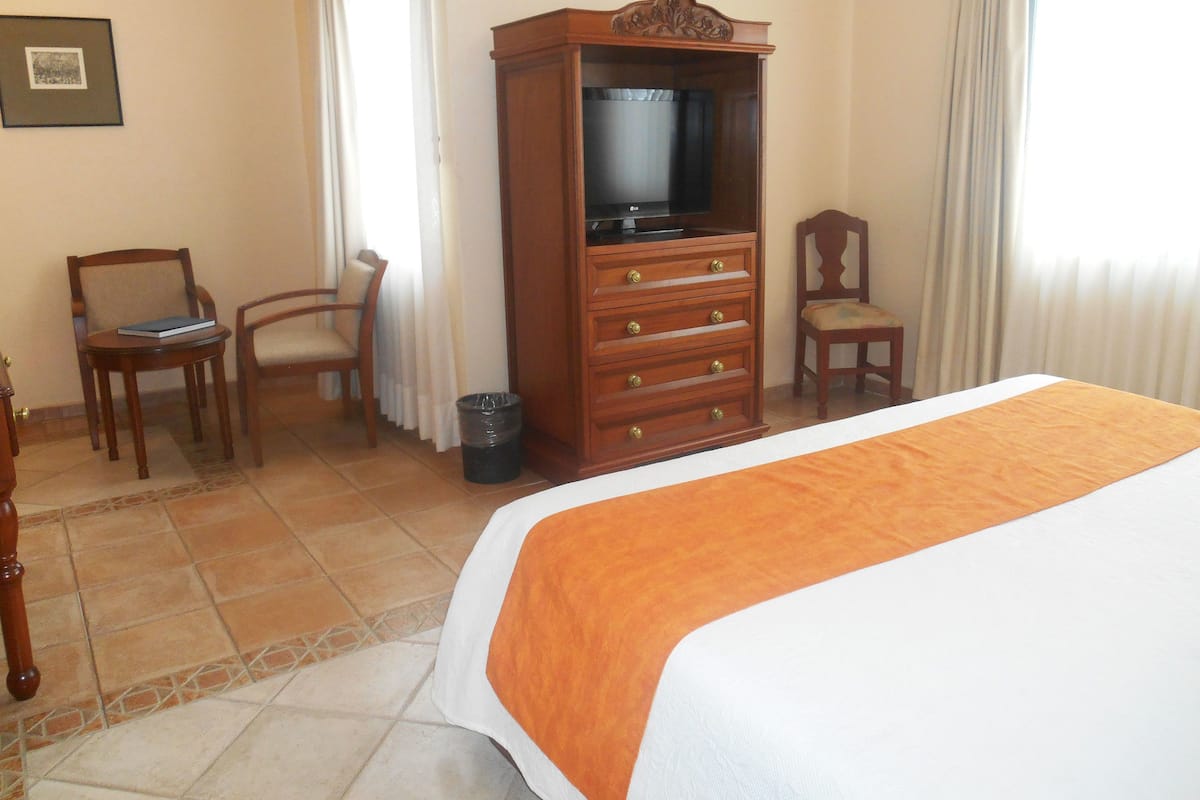 Habitación de hotel con cama, cómoda de madera con cajones, televisor, dos sillas e imagen enmarcada en la pared.