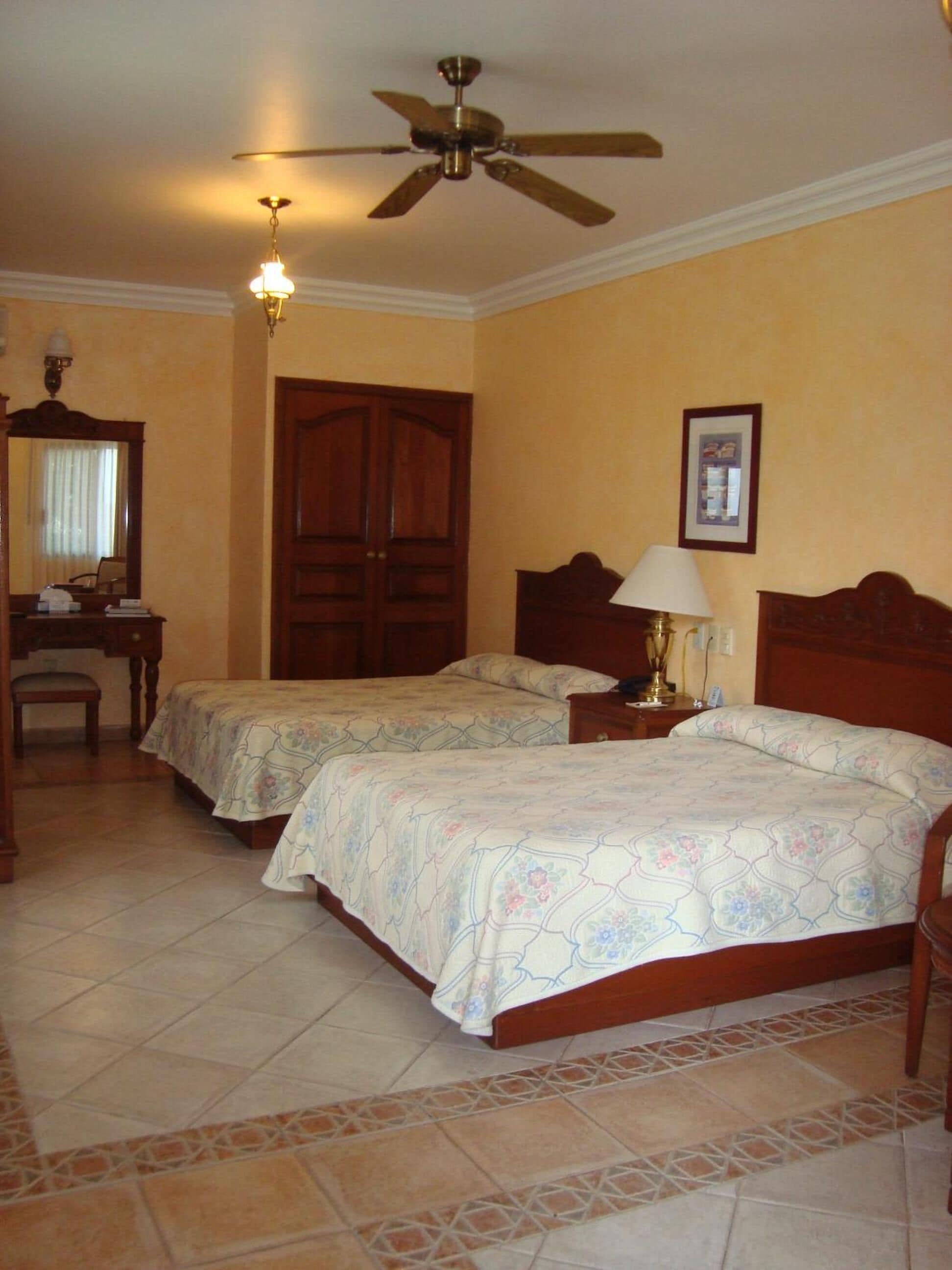 A hotel room with two beds, a wooden wardrobe, a desk with a chair, a ceiling fan, and a framed picture on the wall.