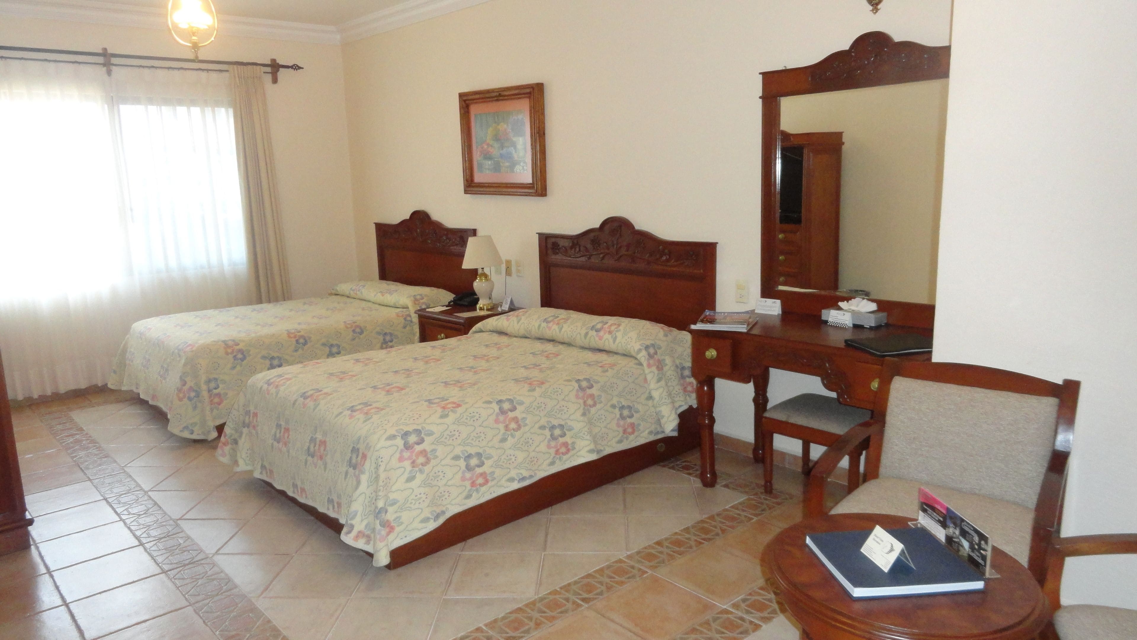 A hotel room with two beds, a desk, a chair, a mirror, and a window with curtains.