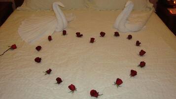 Una cama con una colcha blanca, dos cisnes de tela y un arreglo circular de rosas rojas.