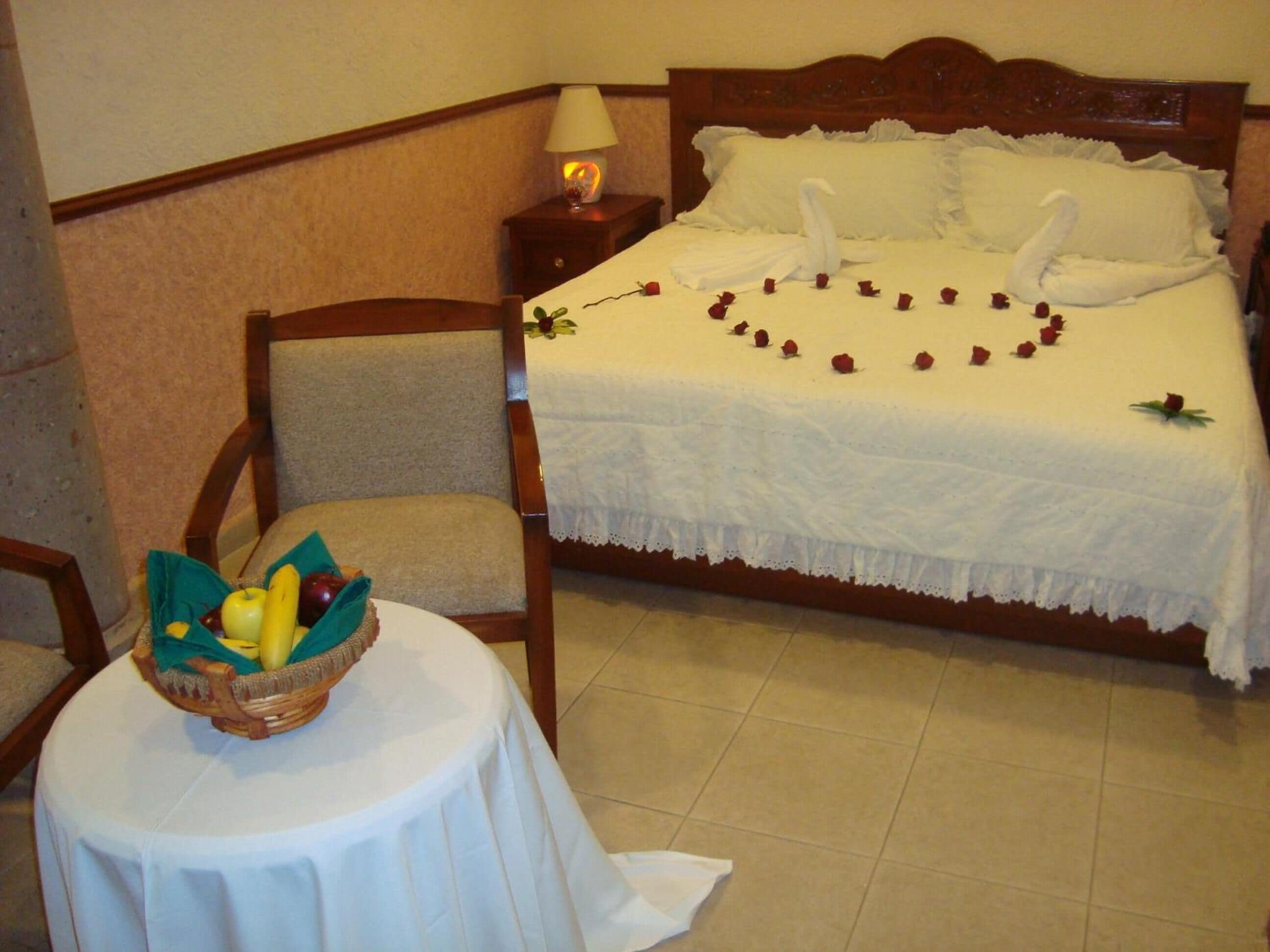 A neatly made bed with white linens and rose petals arranged in a heart shape. A wicker basket with fruits is on a small round table beside the bed.