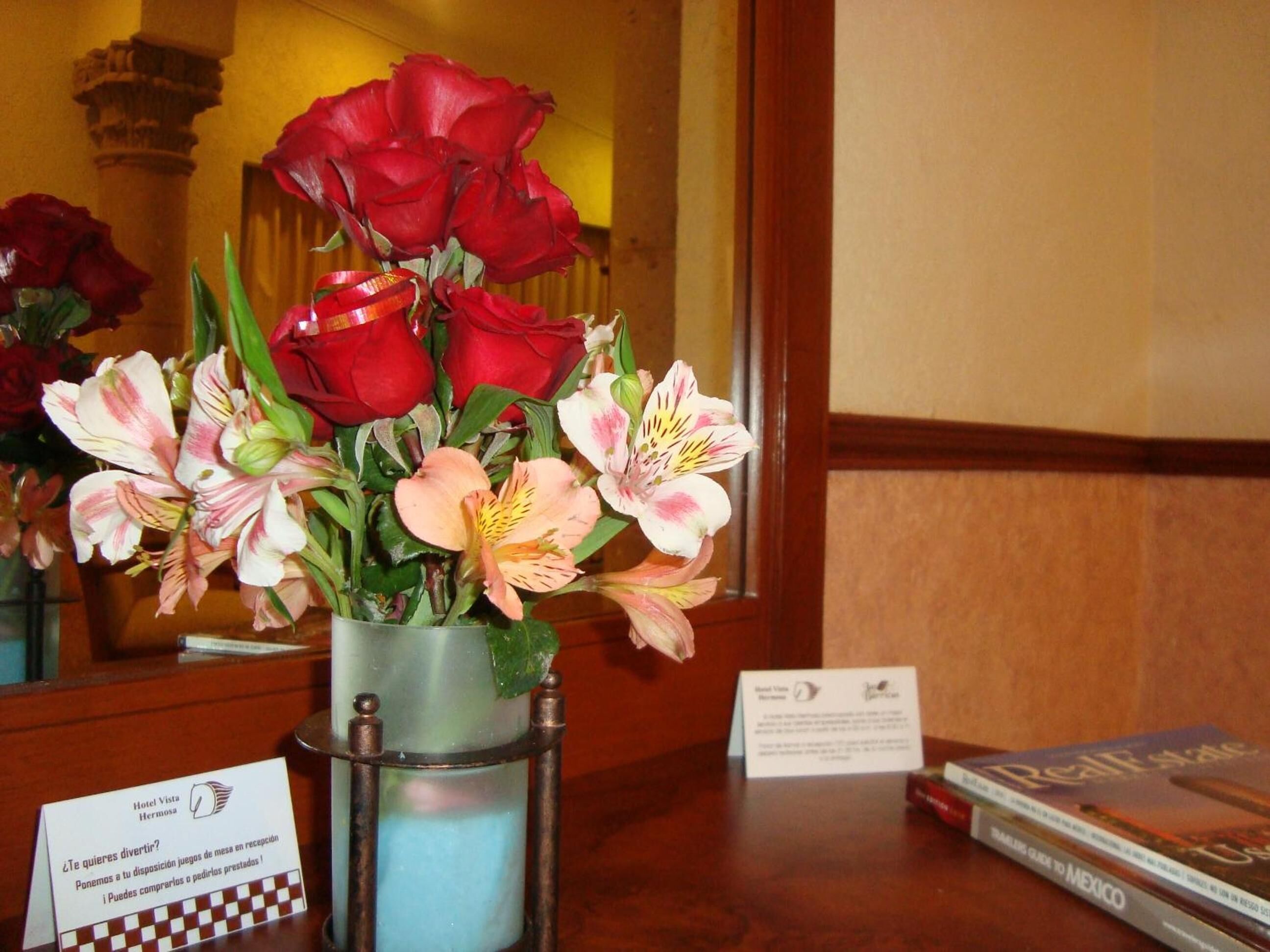 Un escritorio con un jarrón de rosas rojas y flores rosadas y blancas, un folleto del hotel y un cartel con información del hotel.