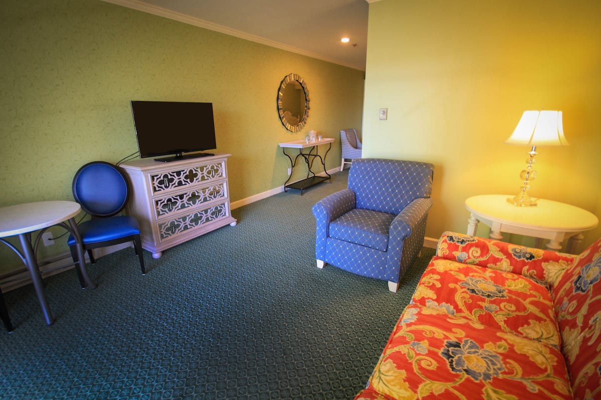 A hotel room with a blue armchair, a patterned sofa, a small table with a lamp, a television on a dresser, and a mirror on the wall.