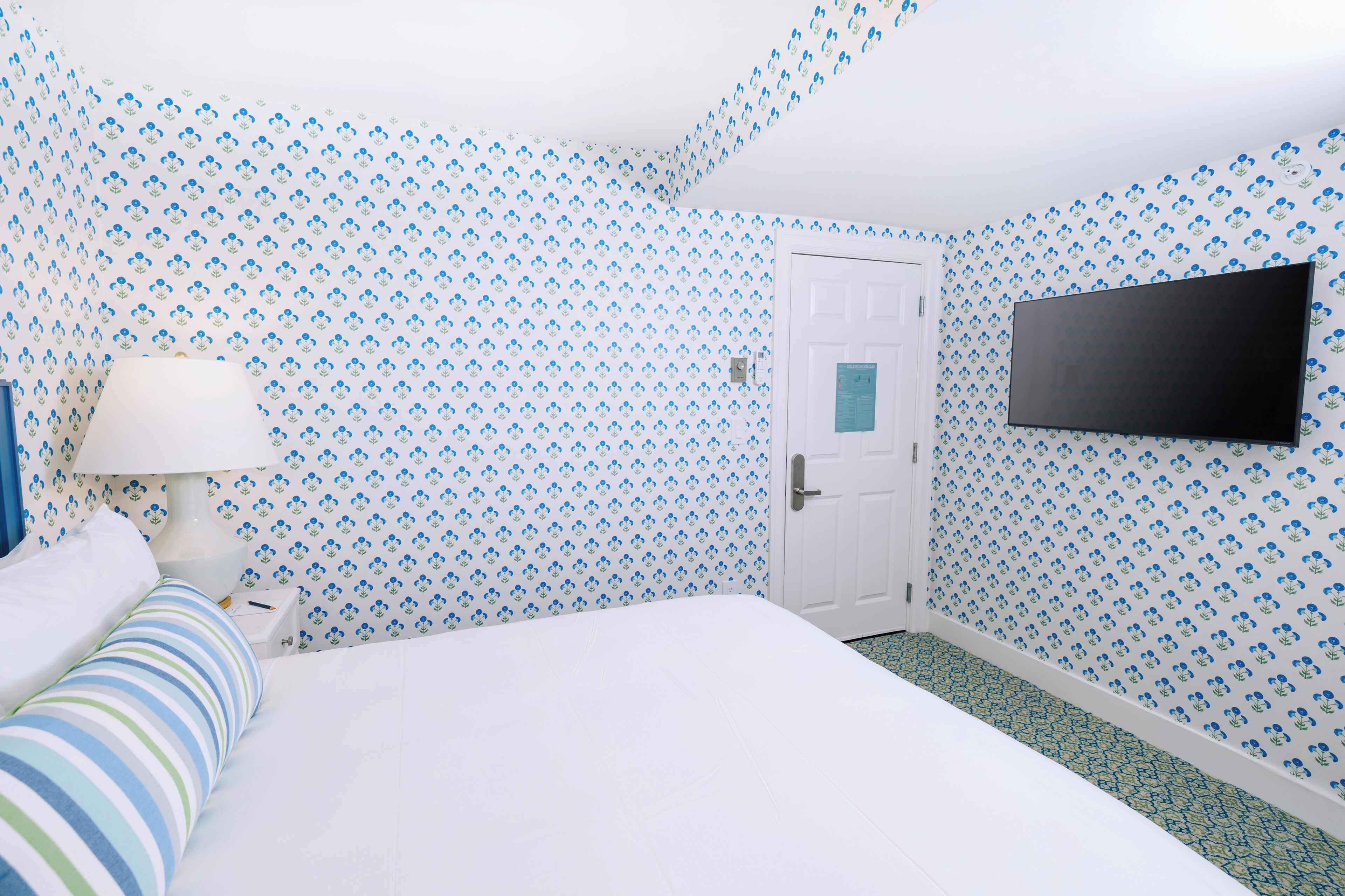 A bedroom with a bed, a TV mounted on the wall, a door, and patterned wallpaper.