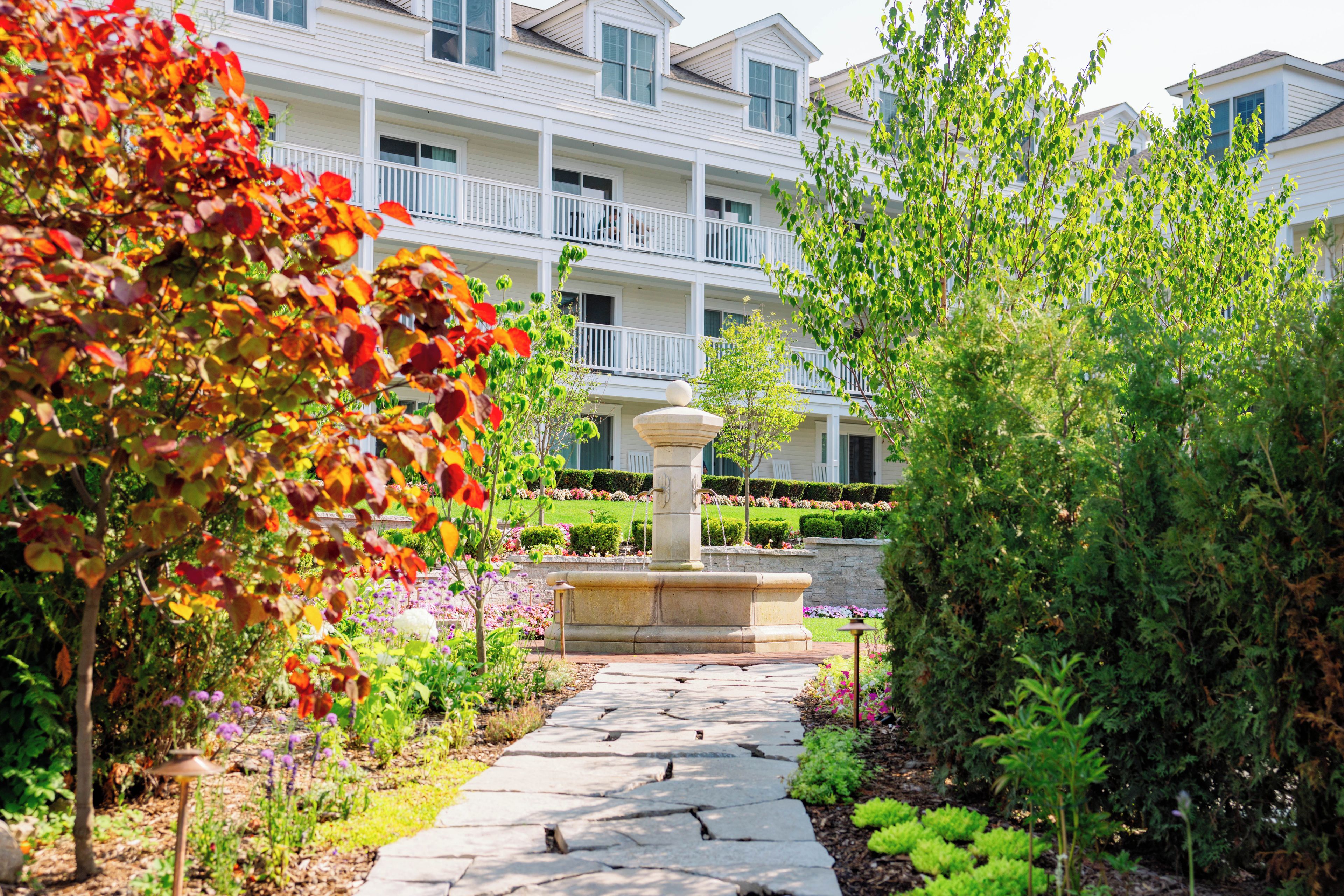 A stone pathway leading to a fountain, surrounded by lush greenery and a multi-story building with balconies.