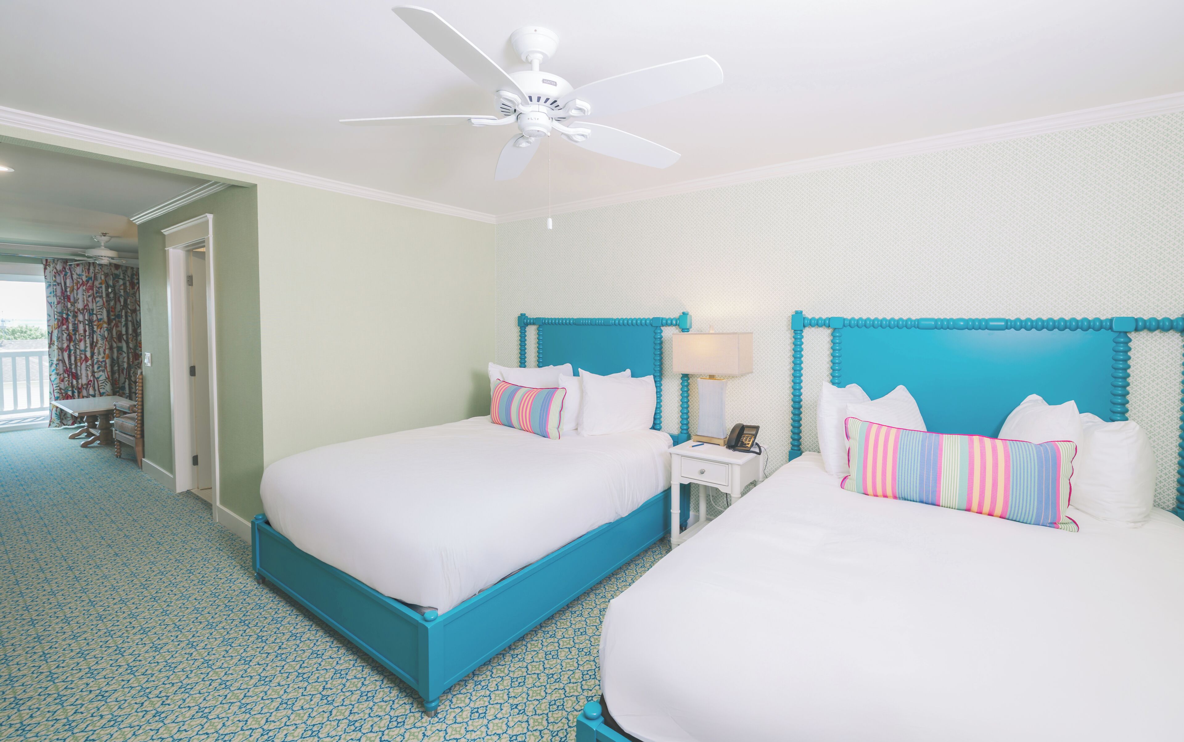 Habitación de hotel con dos camas, ventilador de techo, mesita de noche con lámpara y vista al exterior.