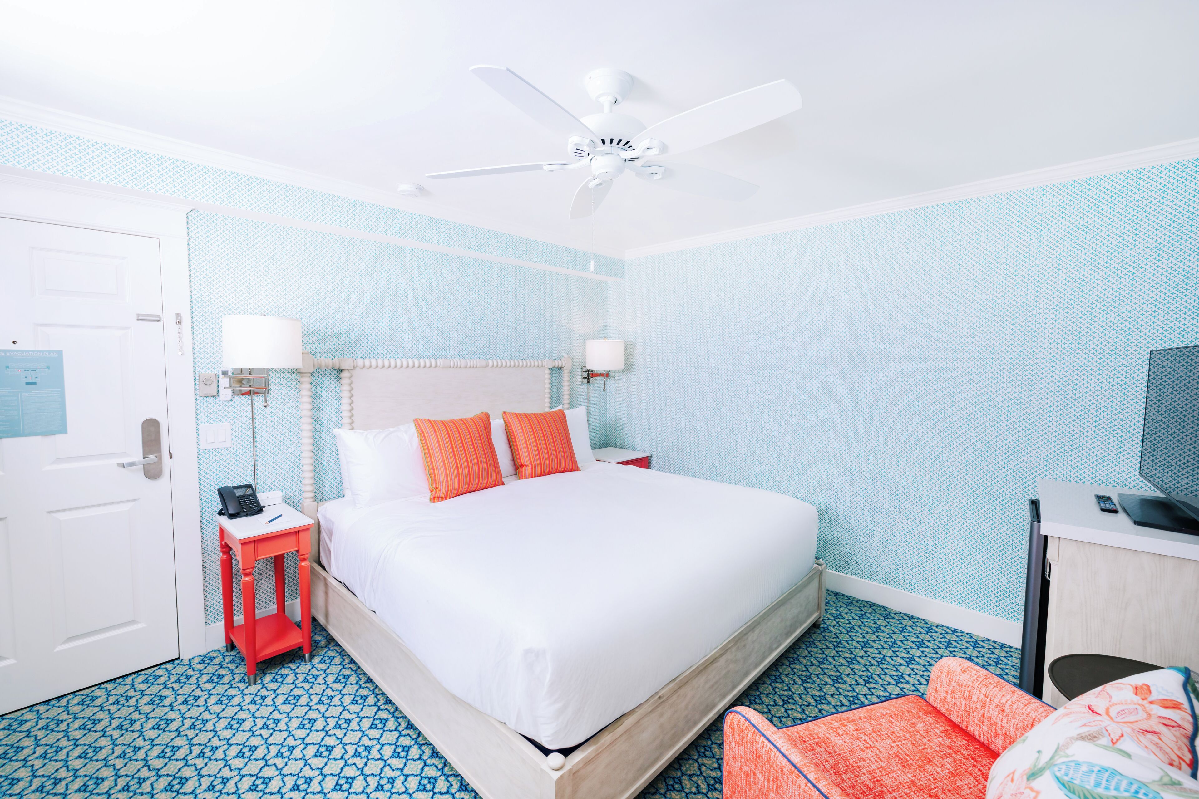 A hotel room with a bed, a desk, a chair, a ceiling fan, and a door.