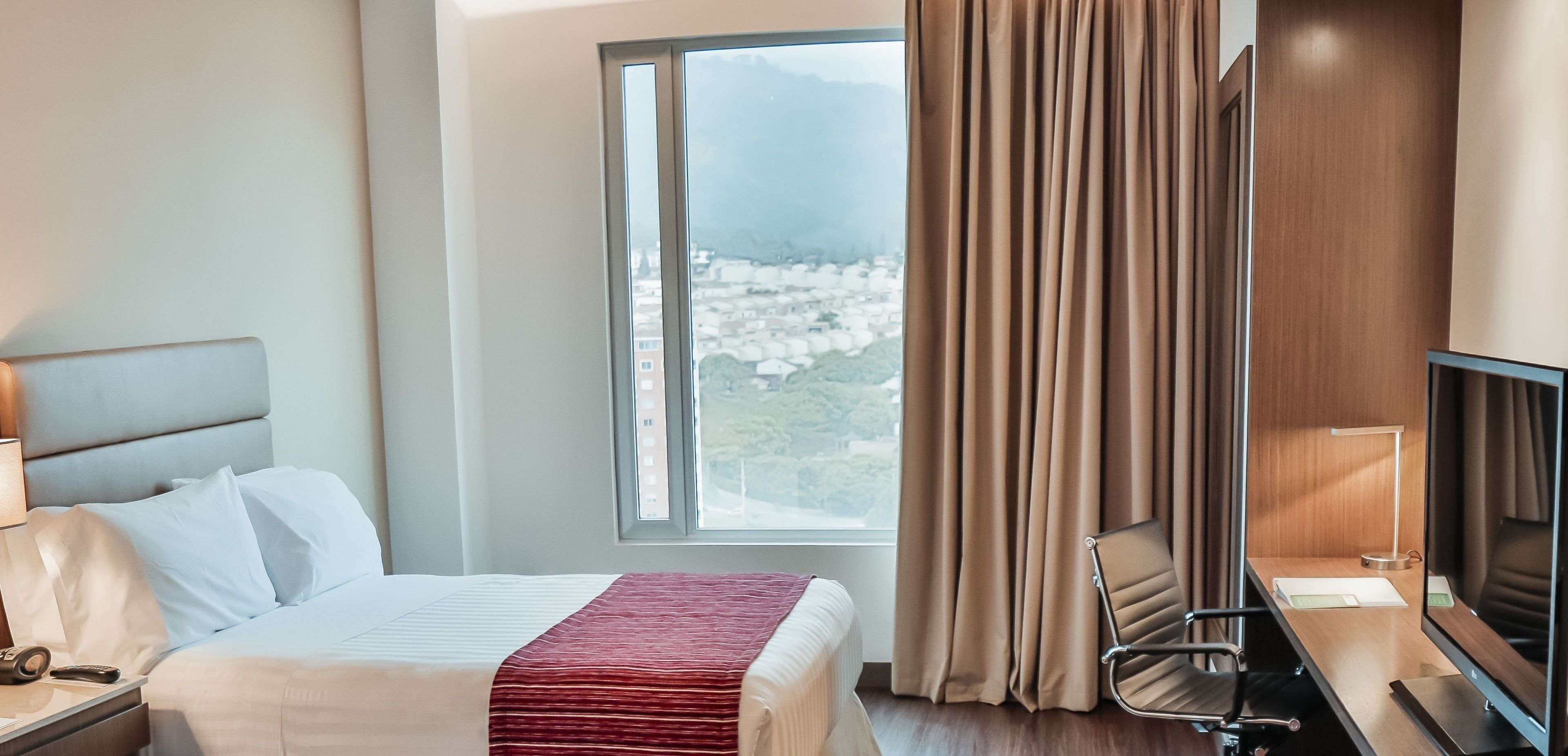 A hotel room with a bed, a desk, a chair, a TV, and a window with curtains.
