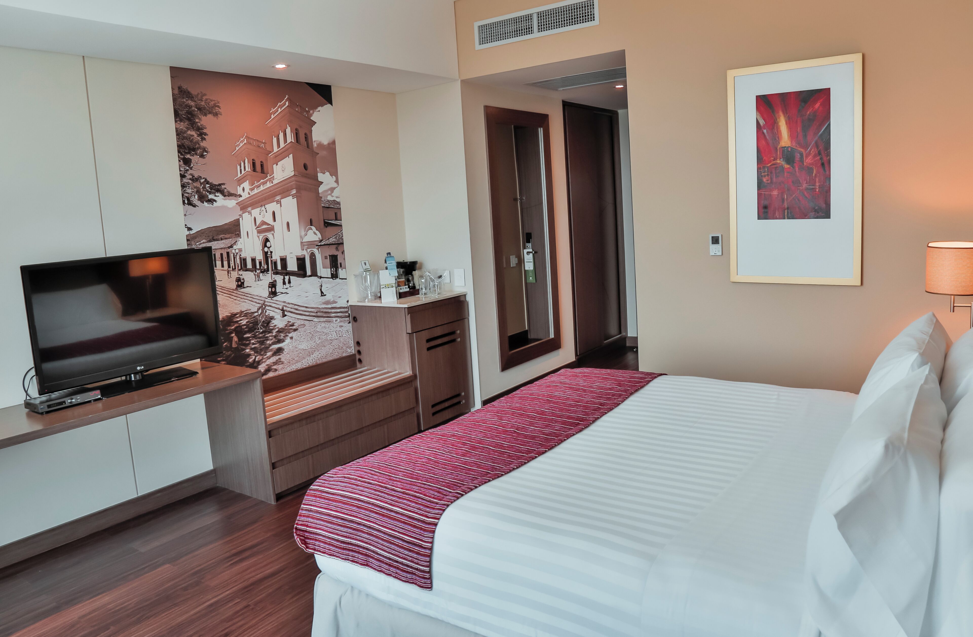 A hotel room with a bed, a TV, a desk with a computer, a mirror, and artwork on the walls.
