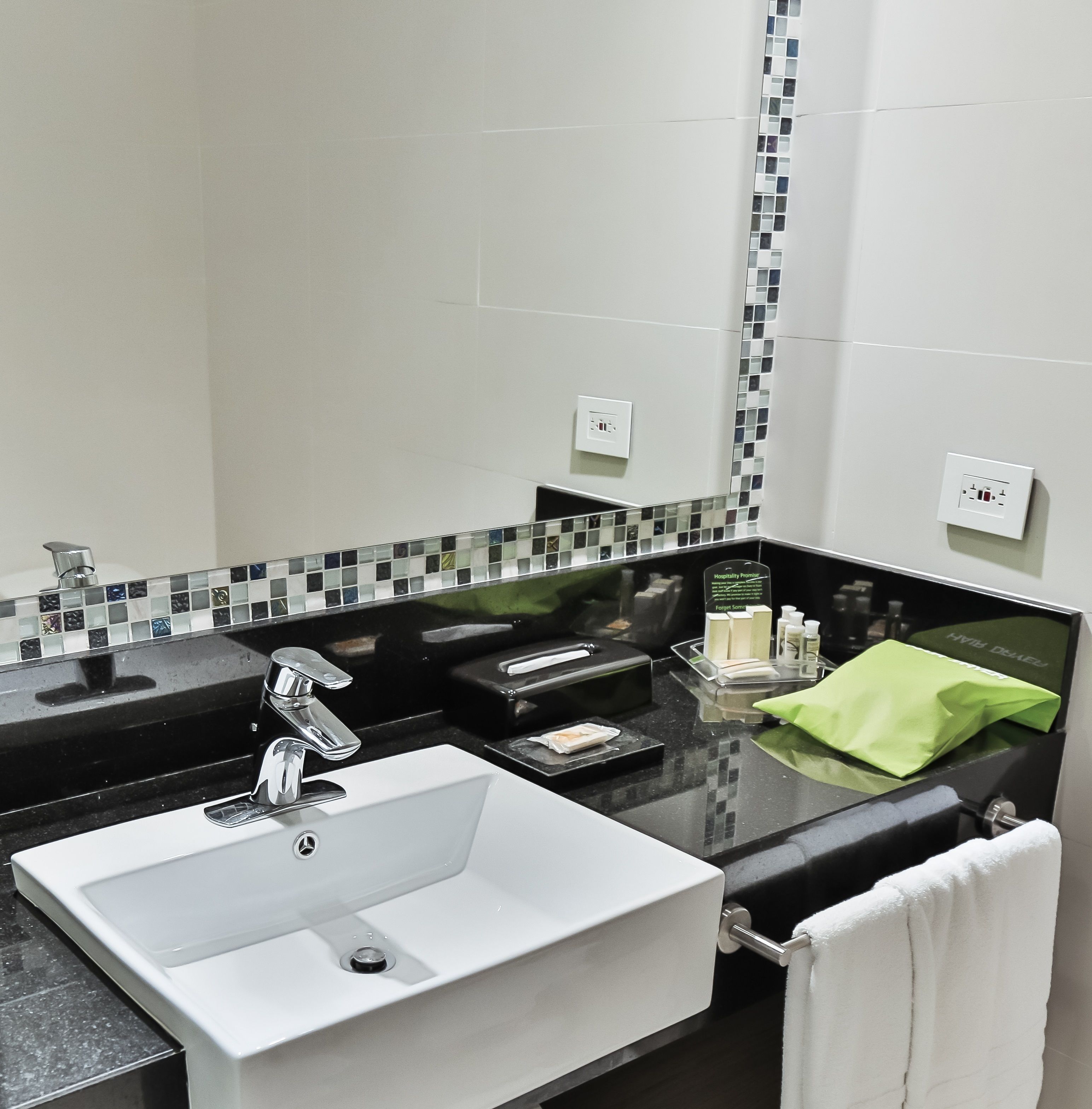 Ein Badezimmer mit weißem Waschbecken, schwarzer Arbeitsplatte und einem Spiegel mit Mosaik-Spritzschutz.